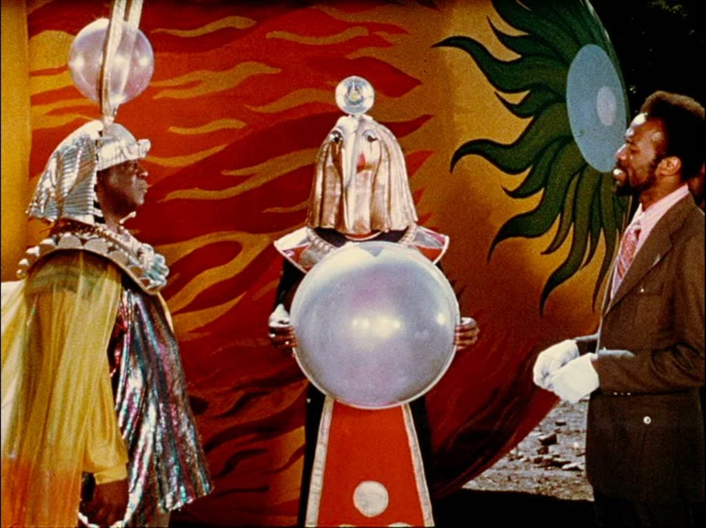 a man in Egyptian garb is facing a godlike creature holding a crystal ball and another in more 1970s attire from a vintage film 