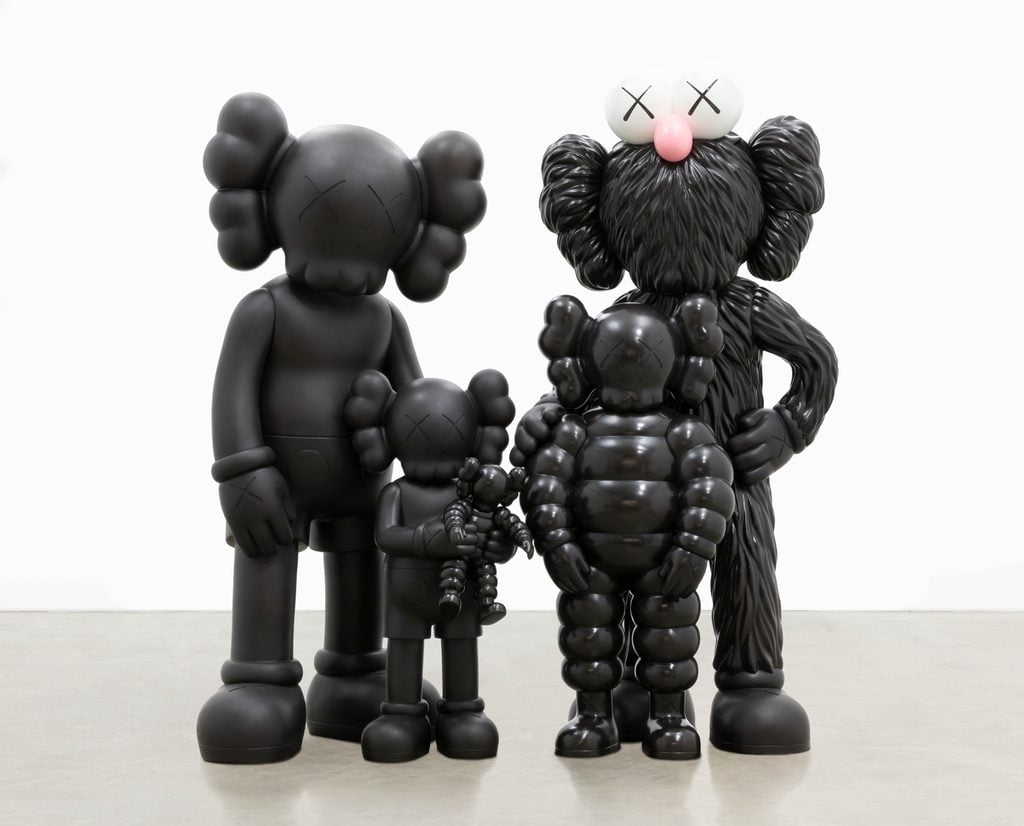 The image shows a family of four black KAWS sculptures. Each figure has distinct shapes and textures: two resemble the iconic "Companion" design with smooth surfaces, while one is a furry character similar to "BFF." The fourth figure appears inflated, akin to "Chum." All figures feature the signature "X" eyes.