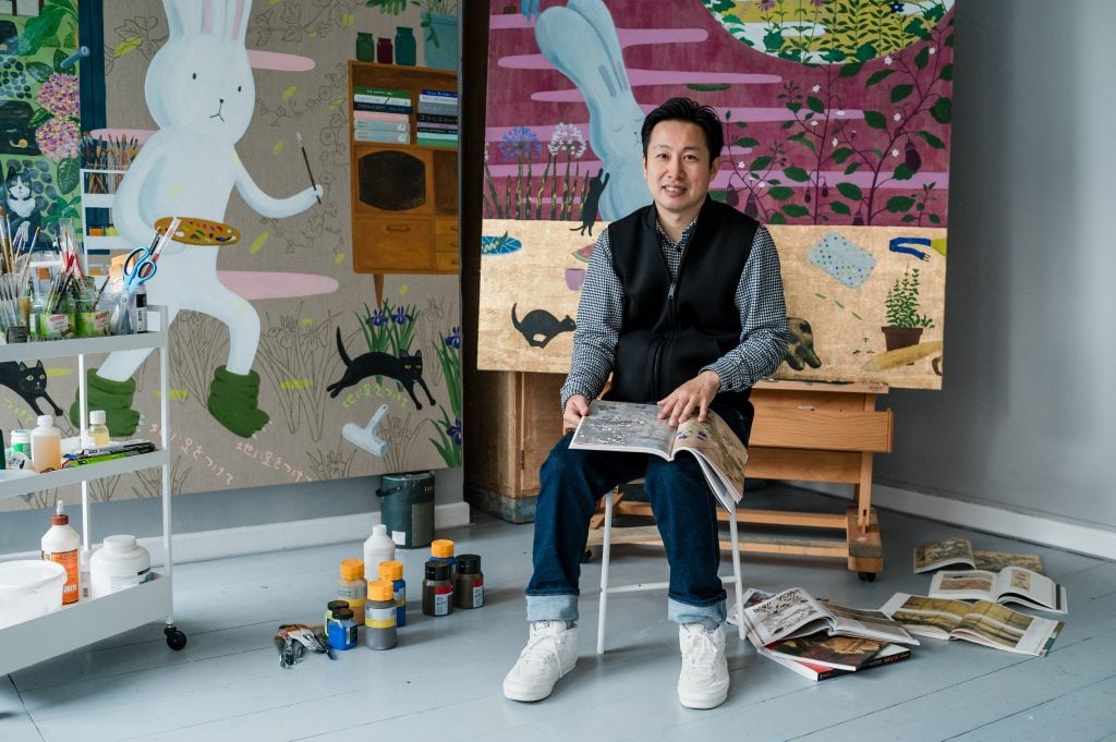 a man sits in a well lit studio surrounded by bright paintings