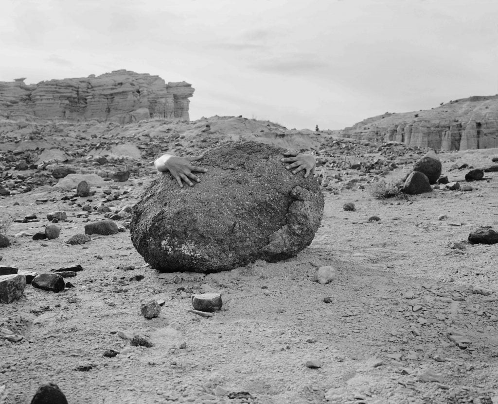 A black and white photograph of the hands of an unpictured person wrapped around a large rock