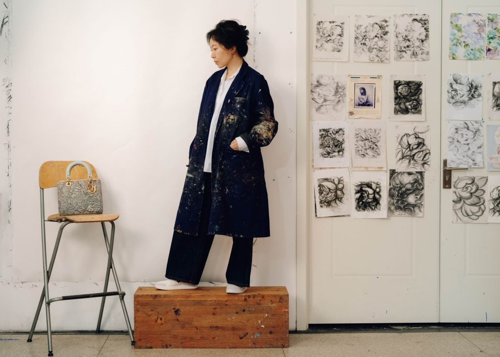 a woman in a black overcoat stands on top of a wooden block and is looking at a purse on a chair 