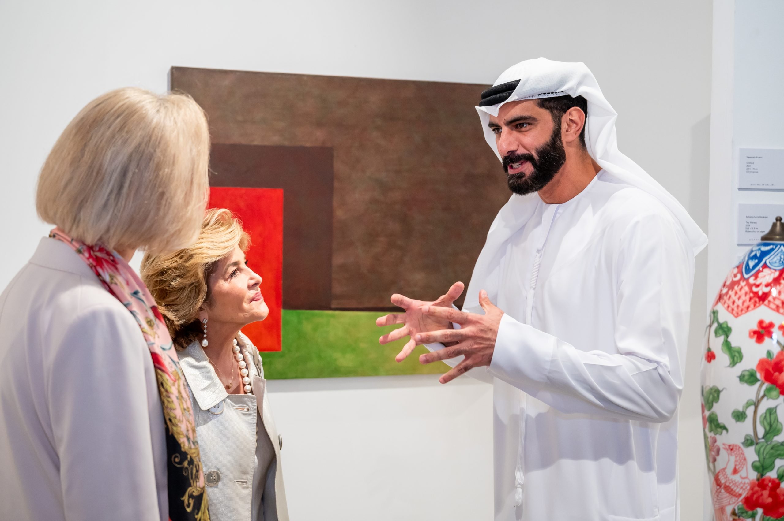 A man in traditional Emirati attire engages in conversation with two women in front of a modern abstract painting at an art exhibition.