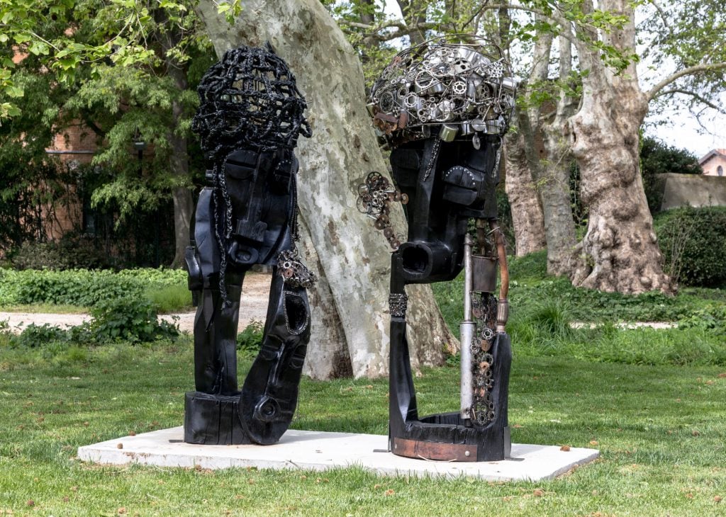 two sculptures in an outdoors garden space, they are vaguely figurative and made of a black material