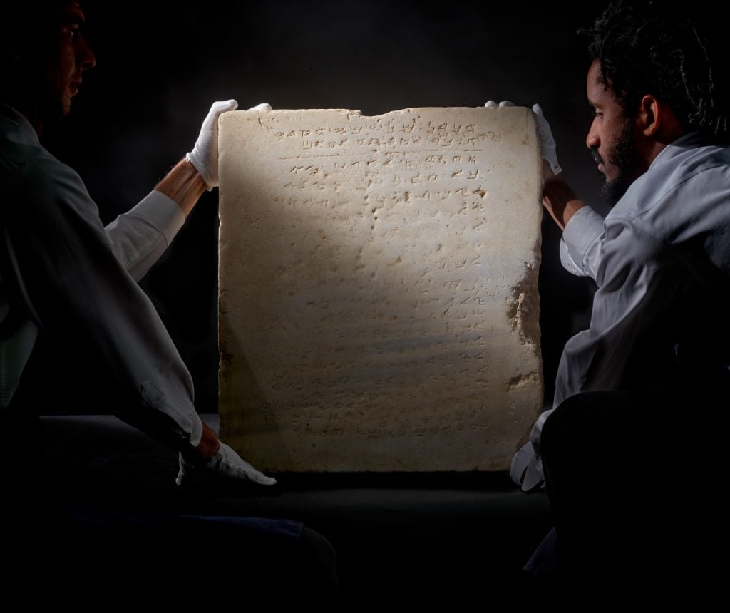 Two workers wearing gloves are seen holding up a stone tablet depicting the Ten Commandments in a dark room.