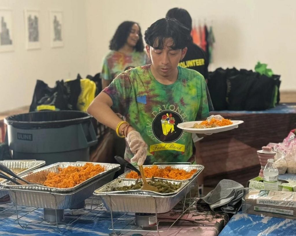 Azikiwe Mohammed's New Davonhaime Food Bank fed 200 people at an event held at Project for Empty Space in Newark as part of the artist's residency there. A man in a green shirt with the food pantry logo is serving food out of an aluminum chafing dish. 