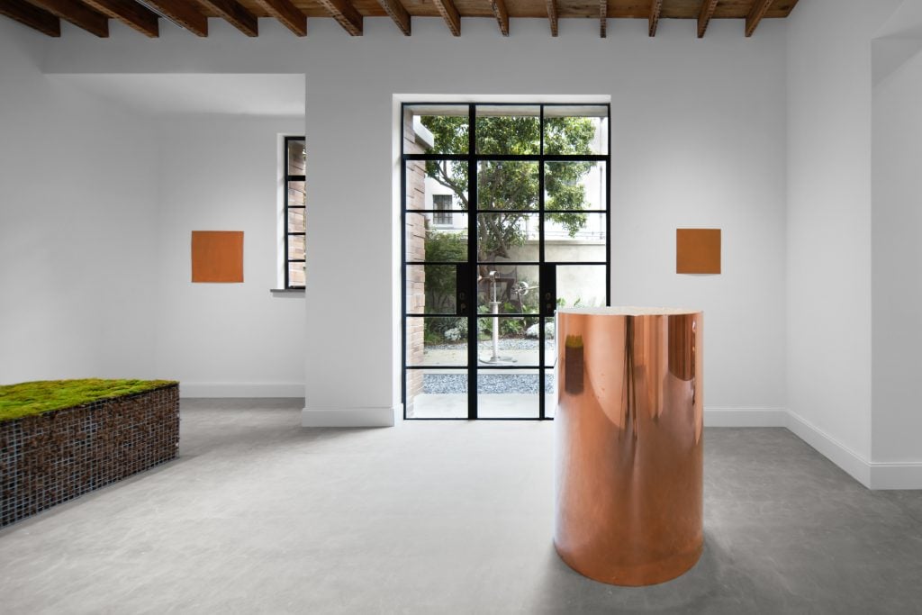 a large copper sculpture stands in the middle of gallery space with a large window overlooking a courtyard in the background