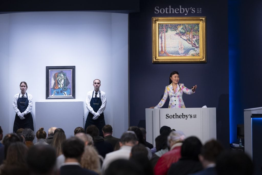 An auctioneer at Sotheby's stands at the rostrum, gesturing during a modern evening auction. Two staff members stand near displayed artworks.