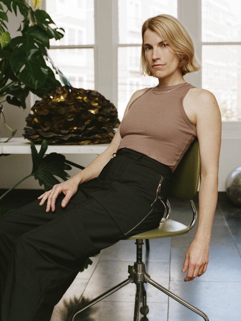 a white woman with golden blonde hair, chin length, sits on a chair in a studio wearing black pants and a sleeveless top