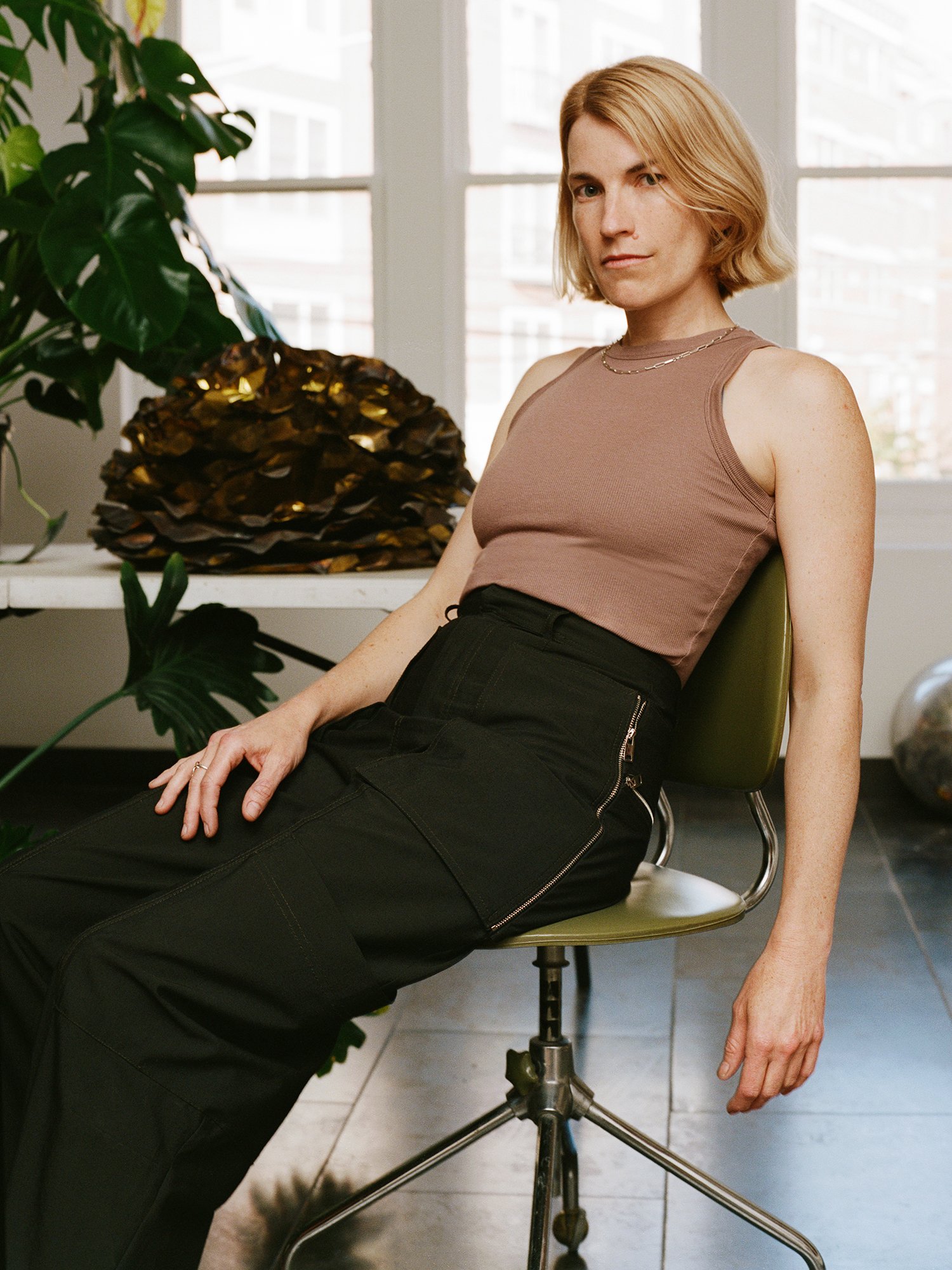 a white woman with golden blonde hair, chin length, sits on a chair in a studio wearing black pants and a sleeveless top