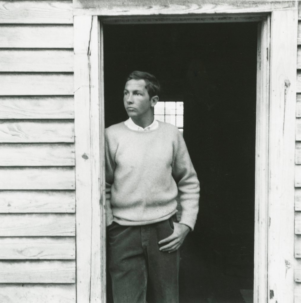 a black and white photo of a young man standing in the open door of a white house