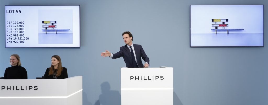 an auctioneer is wearing a suit and has two associates seated next to him and is in mid action 