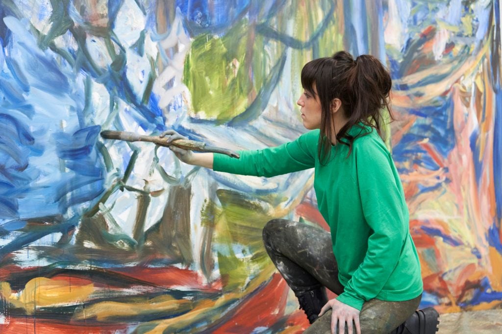 a brunette woman in a green top is seen in profile using a paintbrush to apply paint to a large canvas in tones of blue, white, green, and yellow