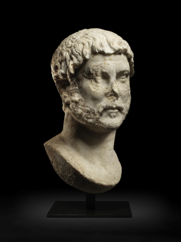 A monumental Roman marble portrait bust of the Emperor Hadrian. The nose and right eye are damaged. The bust is set against a black backdrop.