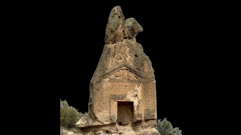 A photograph of Arslan Kaya, a 15-foot tall ancient engraved stone monument, beneath cloudy skies and surrounded by trees.