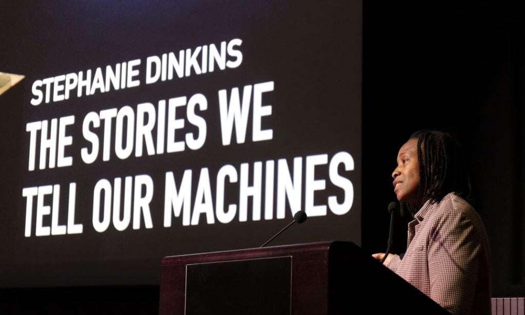 a woman stands at a podium, presumably addressing an audience that is out of view, behind her is a projection of the text 'Stephanie Dinkins The Stories We Tell Our Machines'