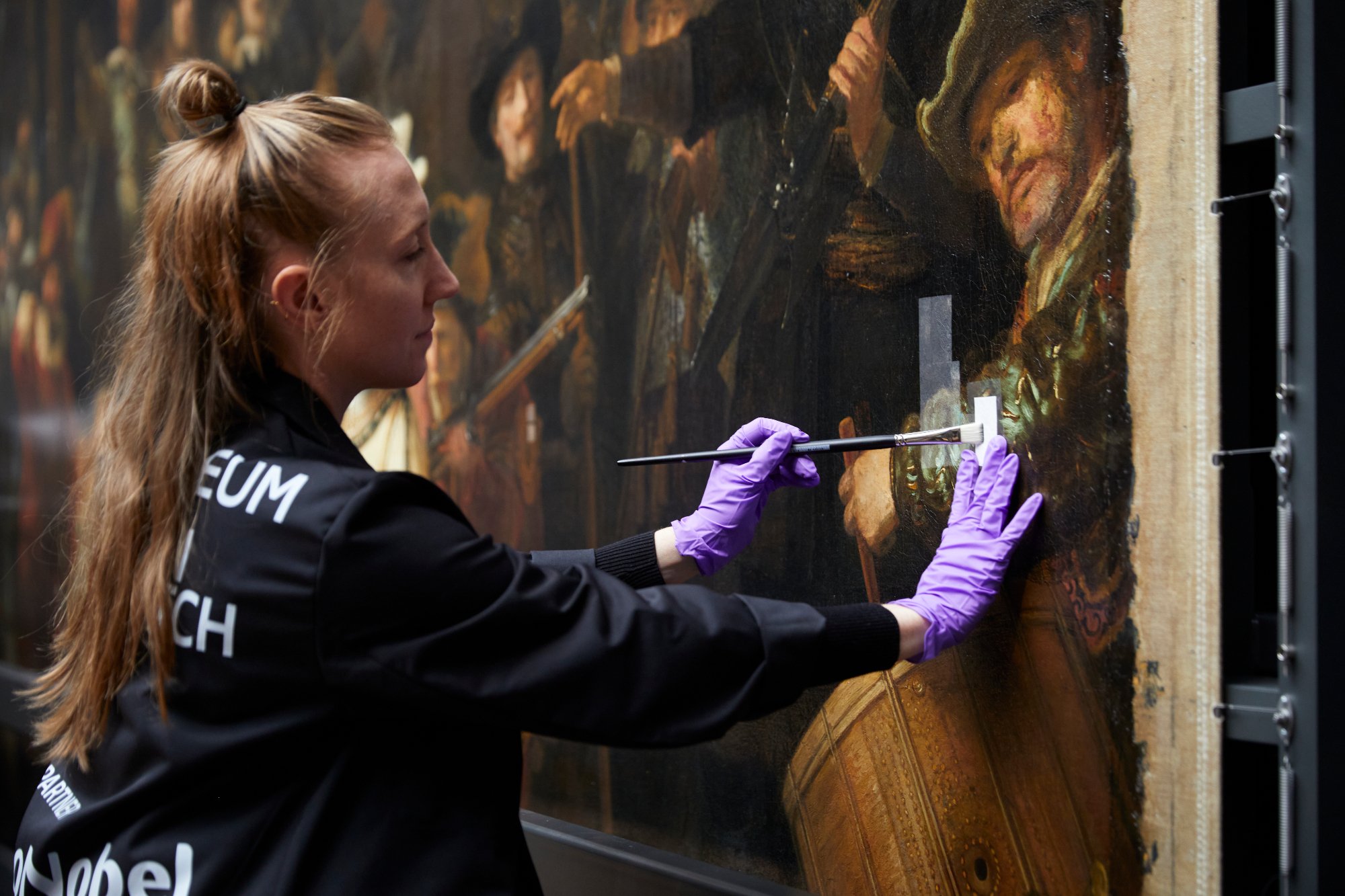 a woman in professional protective clothes and purple protective globes uses a brush to paint some solvent onto a piece of cotton attached to a canvas, she is removing the paintin's protective varnish