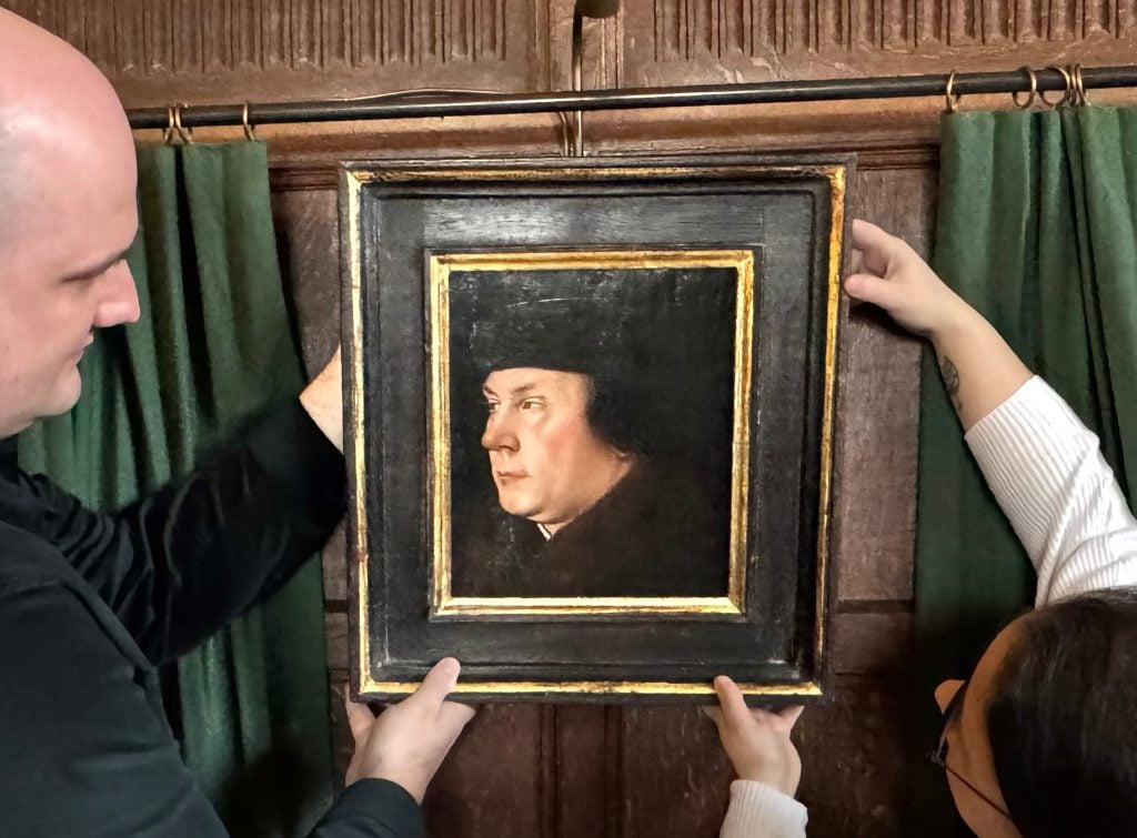 two people hold up a Tudor portrait of a man onto a wood panelled wall with green curtains framing it