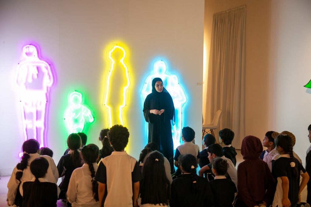 Abu Dhabi Art docent leads a group of children through an installation of neon outlines of people in different colors.