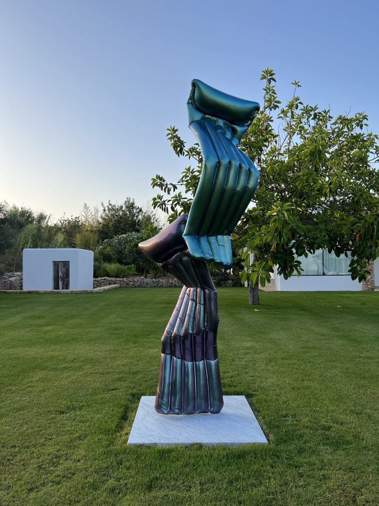 A metallic, large-scale sculpture installed on a green lawn by Adam Parker with two elements resembling pool floats, bent and stacked on top of one another.