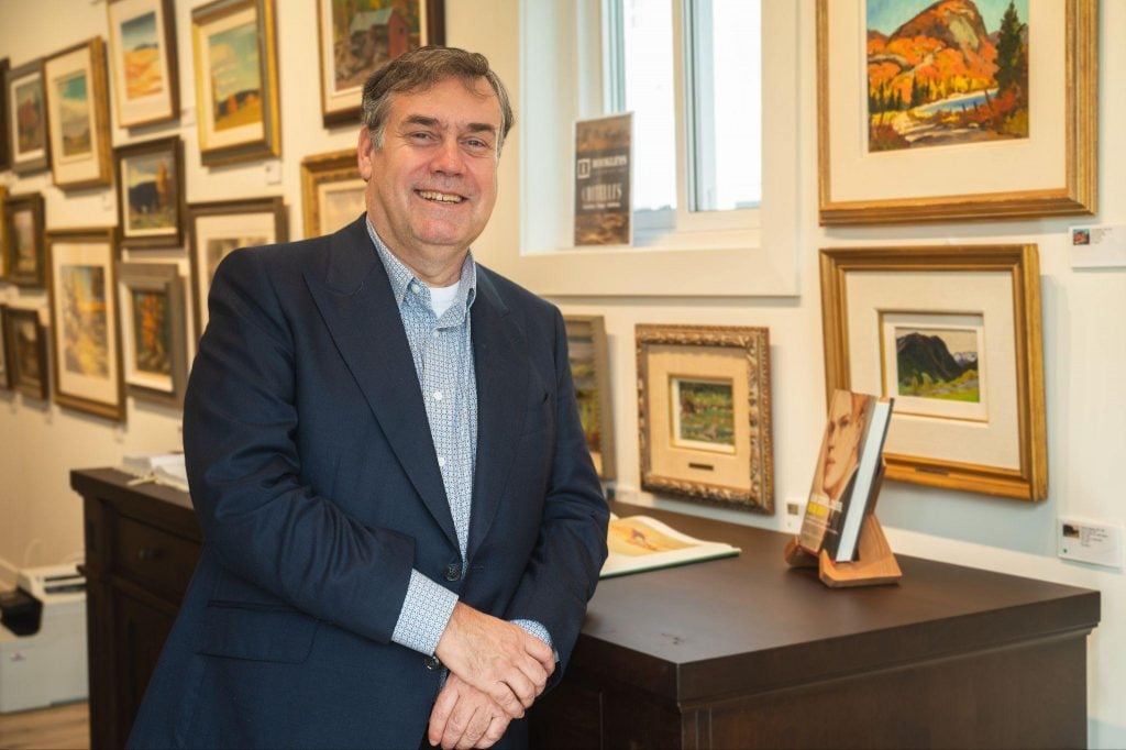 Portrait of gallerist Andrew Rookley wearing a navy suit and ligh blue button down seated on a desk with a wall of small-scale artworks in gold frames covering the wall behind him.