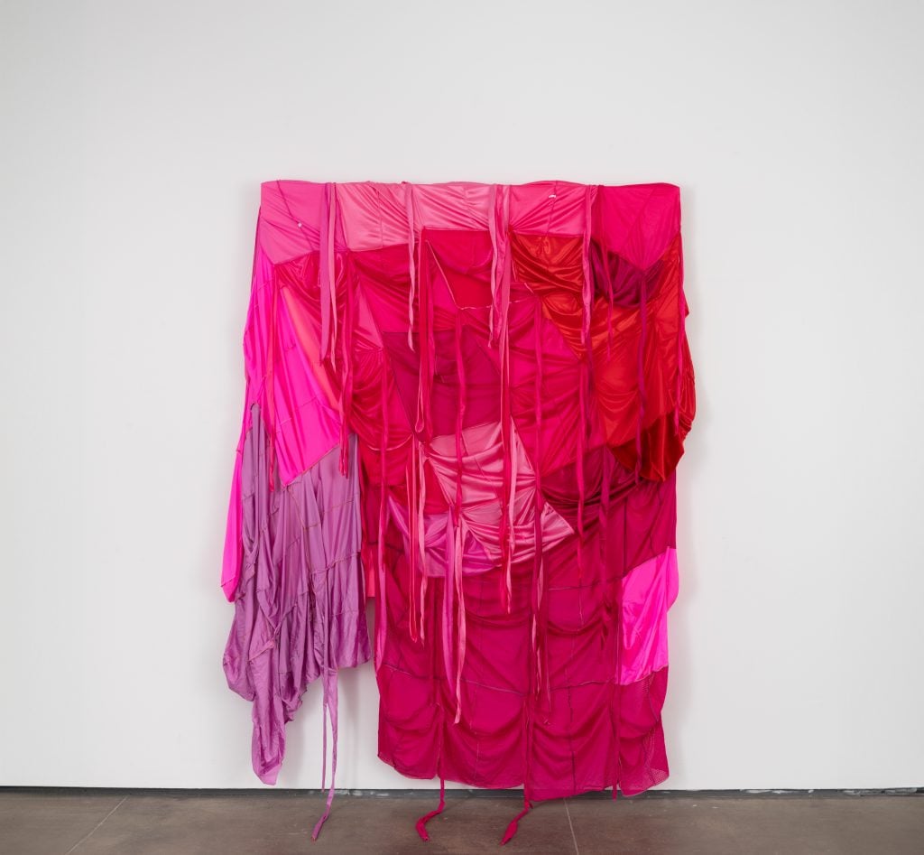 A sculpture made of pink fabric hangs from a white gallery wall