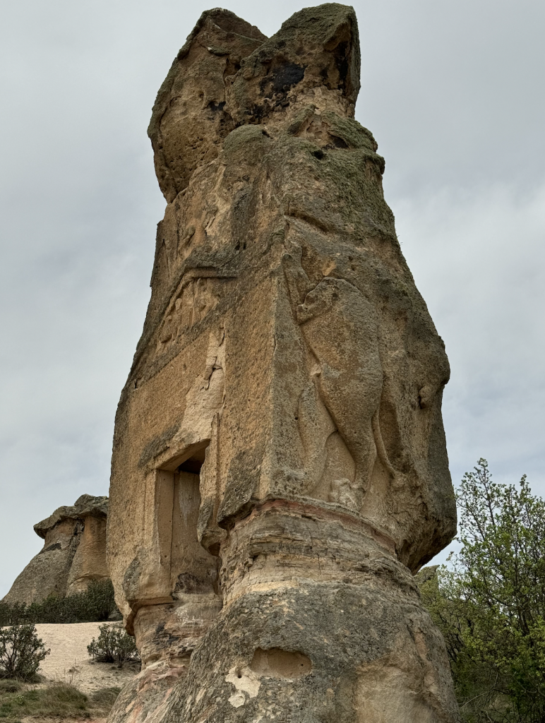 A photograph of Arslan Kaya, a 15-foot tall ancient engraved stone monument, beneath cloudy skies and surrounded by trees.