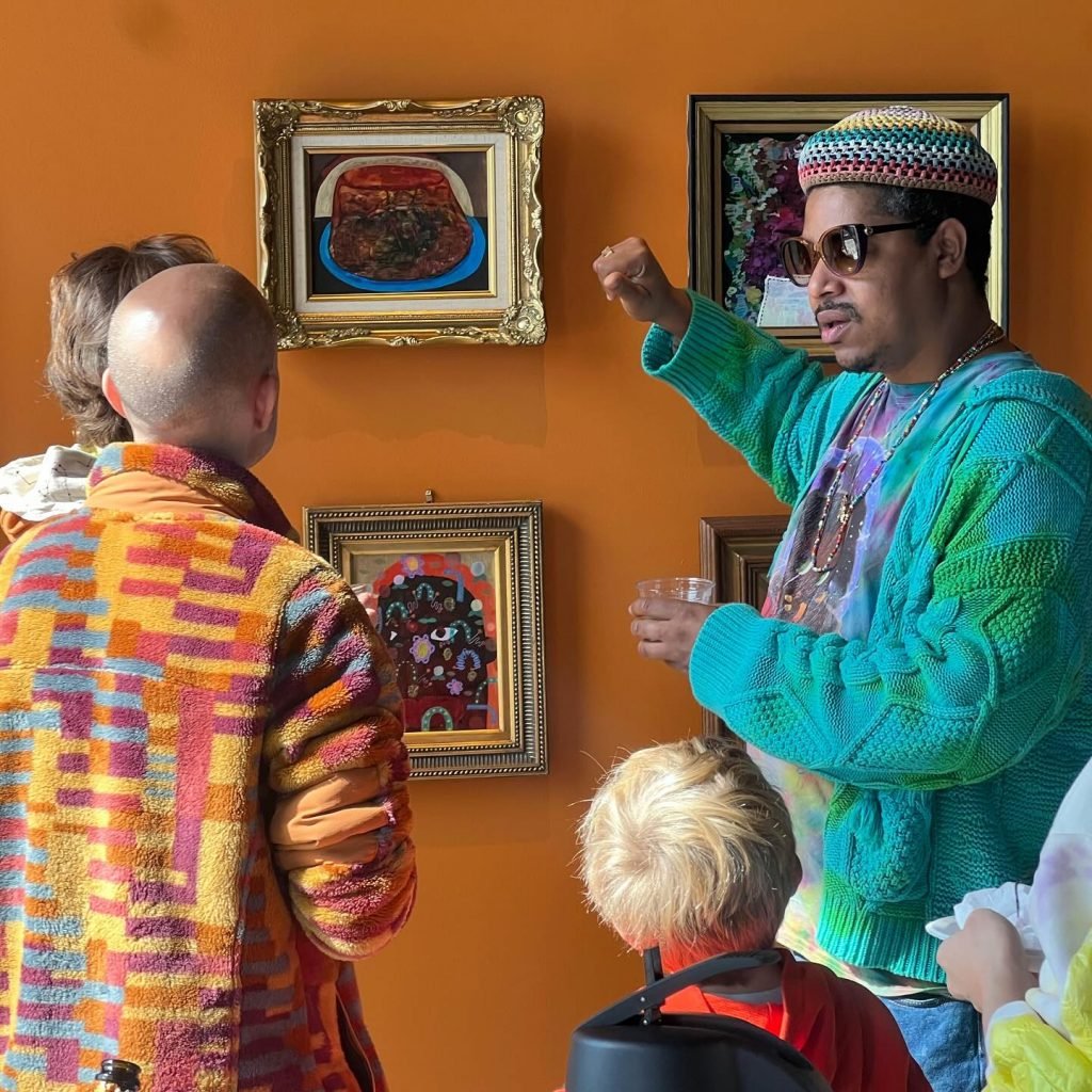 Azikiwe Mohammed speaks with visitors at his show "Trains, Buses, and the Four C’s" at Project for Empty Space Ironside, Newark. The artist is wearing sunglasses, a bright blue and green cable knit cardigan over a colorful t-shirt, a beaded necklace, and a colorful knit beret. The walls are painted orange and the artworks are stylized still life and portrait paintings of a fairly small size in gold frames the artist sourced from thrift stores.