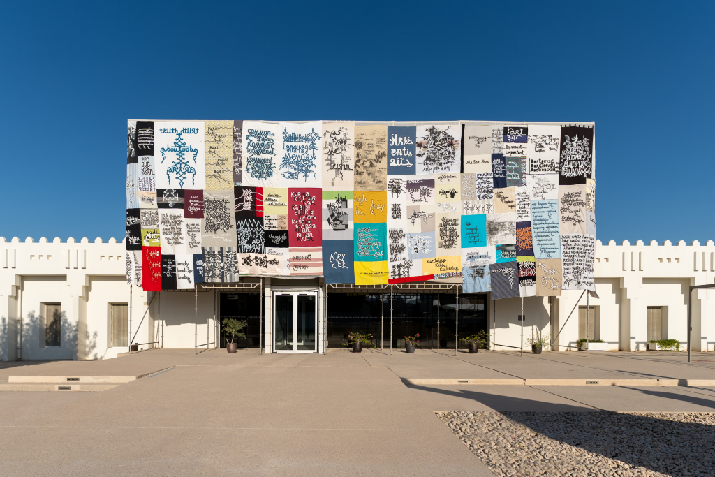 Exterior facade view of Mathaf: Arab Museum of Modern Art, Doha, covered in stitched together, variously colored banners with various Arabic text written on each.