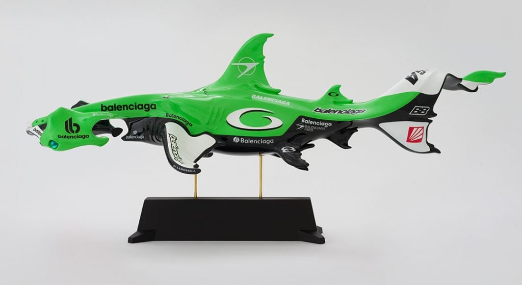 A photograph of a green sculpture depicting a Hammerhead shark using car parts.