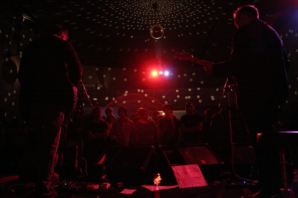 A photograph of a crowd gathered for a concert in a dark room illuminated with red light reflecting off a disco ball.