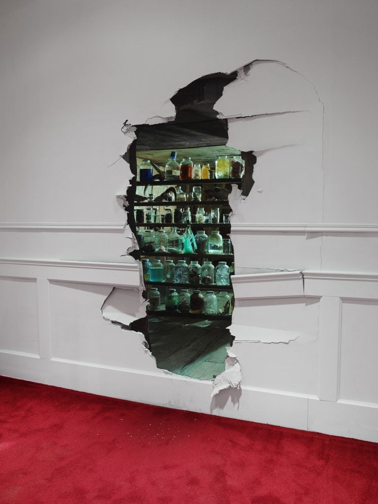 A photograph of a white wall with a hole broken into it leading into a green-lit space, with a red carpet before it.