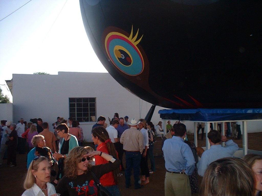 A photograph of a black baloon printed with eyeballs looming over a crowded outdoor courtyard during daytime.