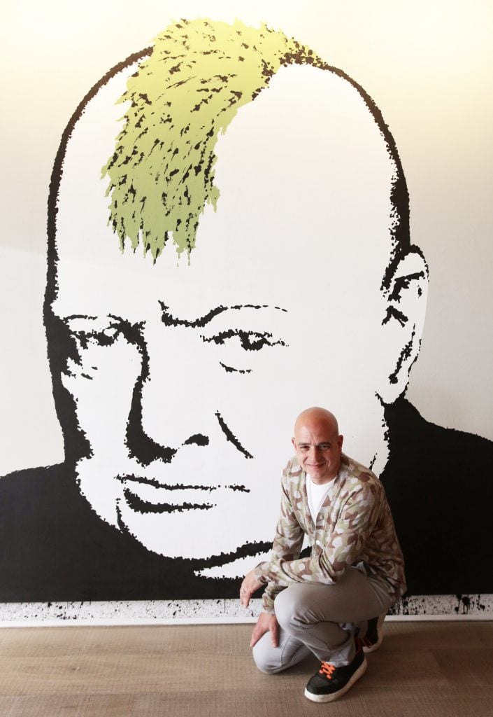 Steve Lazarides poses in front of a large painting of Winston Churchill with a mohawk haircut