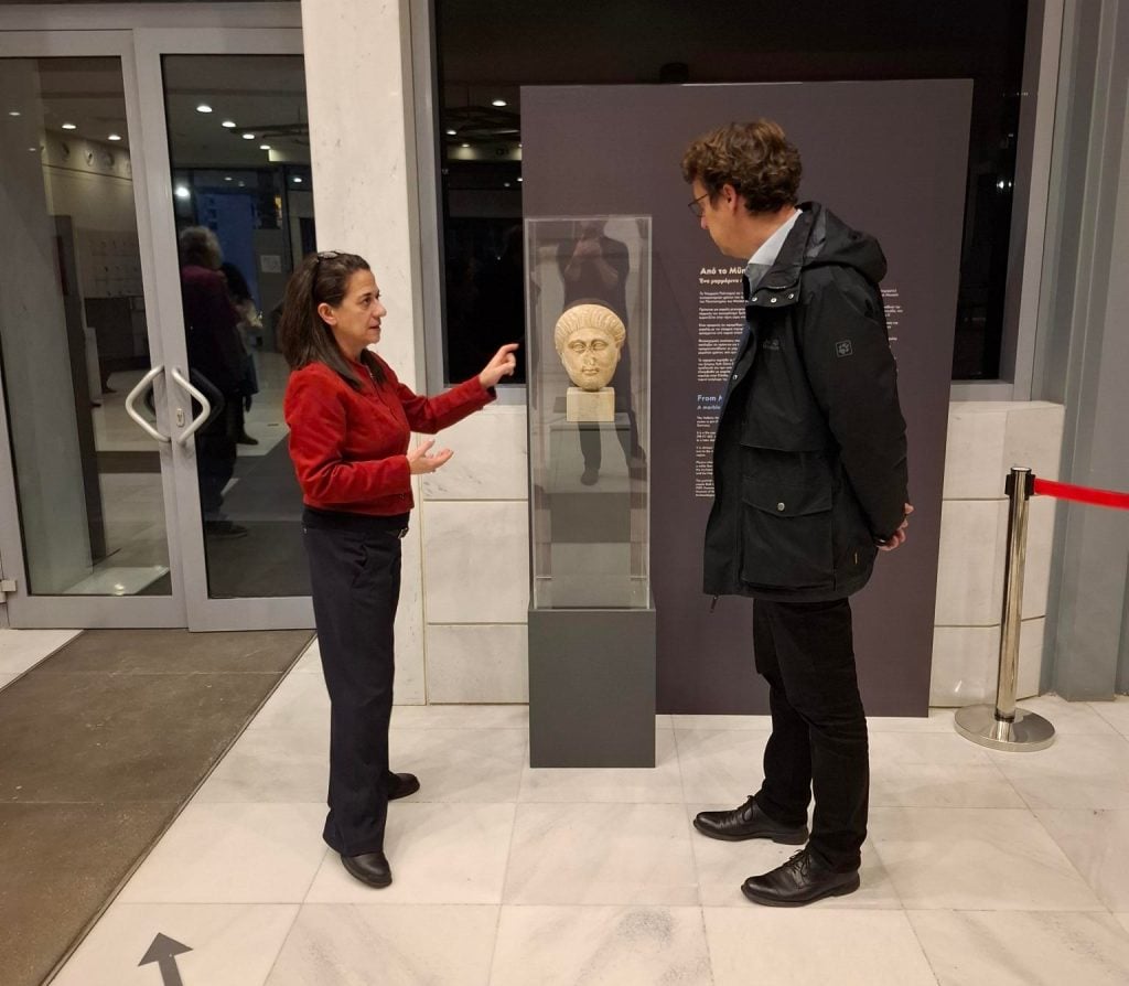 two people, a man and a woman, stand in an interior space like a museum and gesture at a marble head inside a glass case