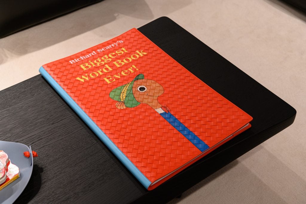 A photograph depicting a Bottega Veneta Intrecciato woven copy of the Biggest Word Book Ever by Richard Scarry sitting on a black table atop a white carpet.