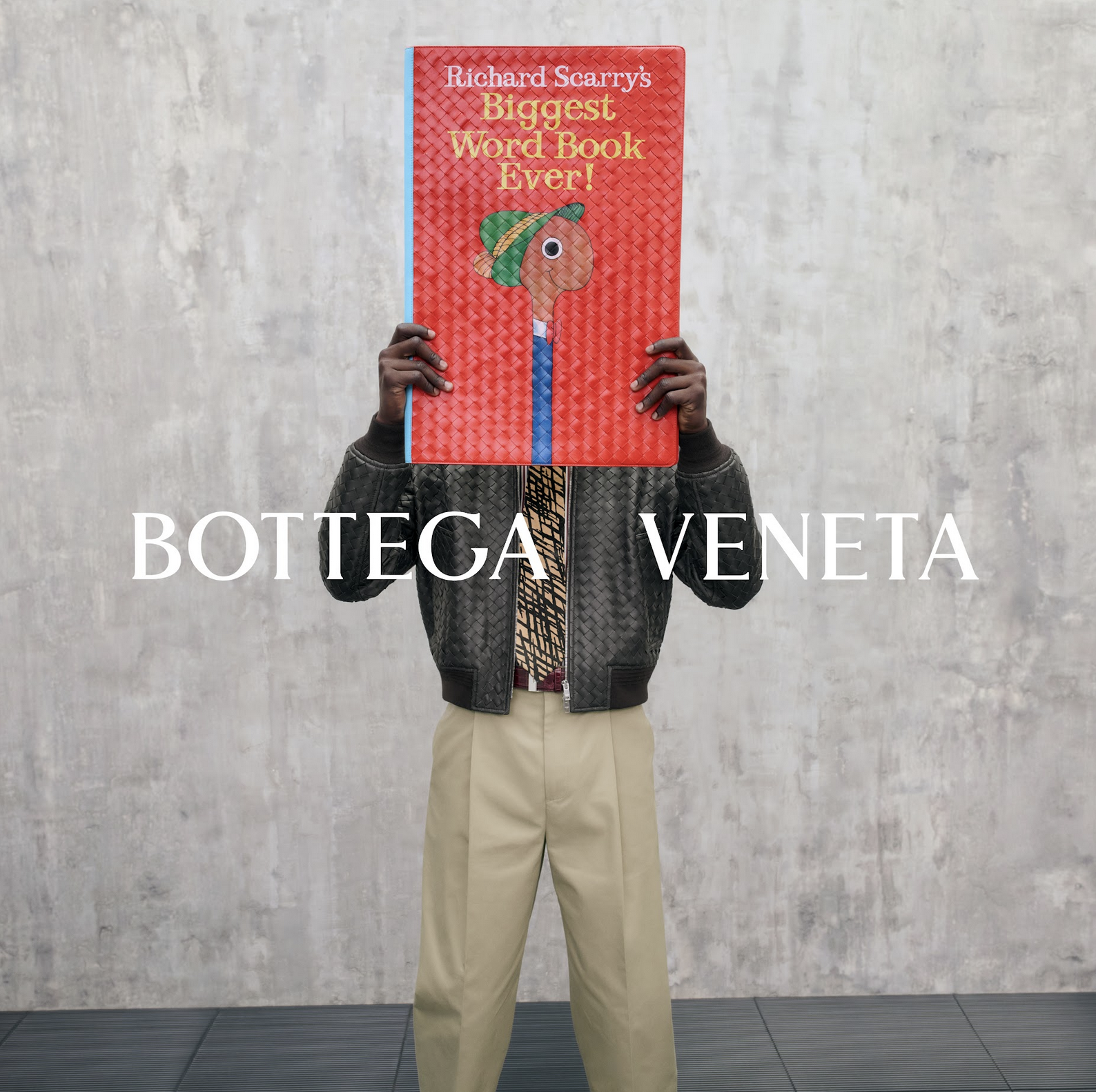 A Bottega Veneta advertisement featuring a well dressed man holding a copy of the house's new Biggest Word Book Ever by Richard Scarry in front of his face, before a grey wall, with the brand's name overlaid atop it all in white.