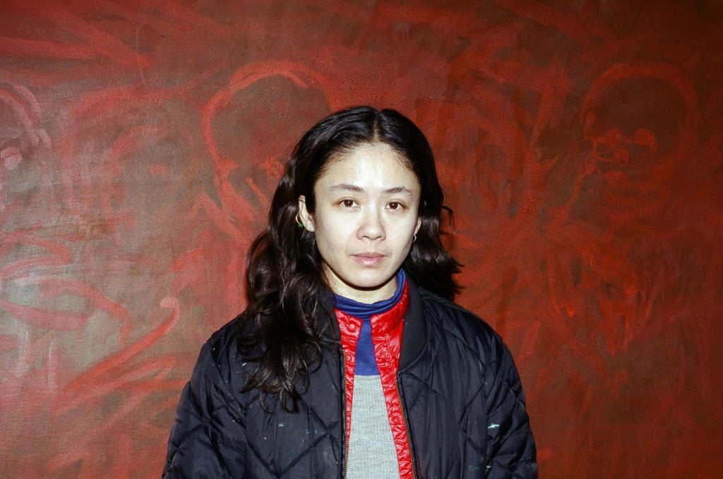 a photo of a 30-something asian woman in a bomber jacket standing in from of a red painting