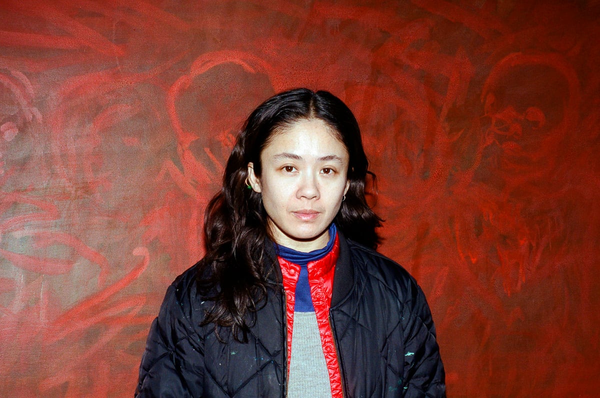 a photo of a 30-something asian woman in a bomber jacket standing in from of a red painting