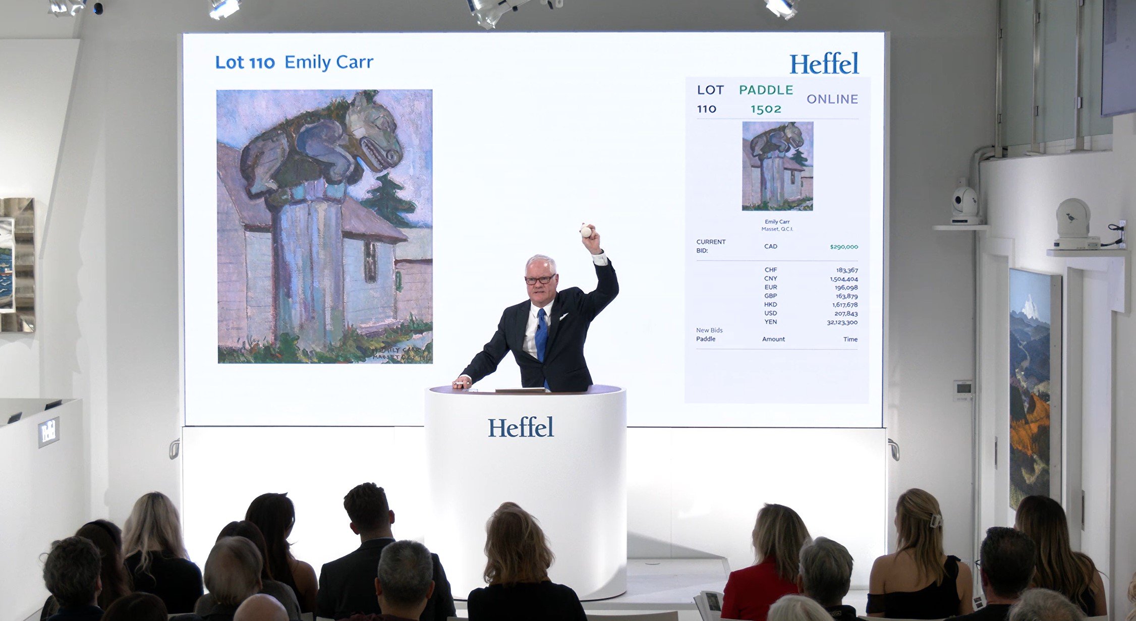 A photograph of an auctioneer standing behind a white Heffel podium with his arm raised before a crowd with the Emily Carr work up for sale projected behind him.