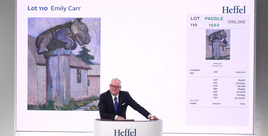 A photograph of an auctioneer at a white Heffel podium with the Emily Carr work up for sale projected behind him.