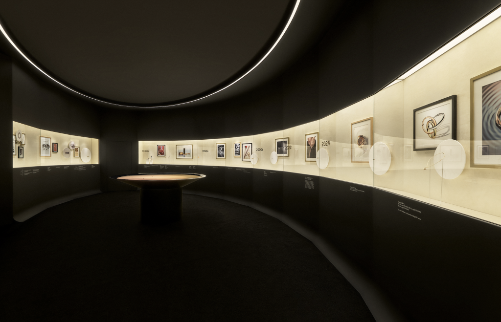 A photograph of a dark, round exhibition space with a brightly lit, curved case highlighting the evolution of the Cartier Trinity ring.