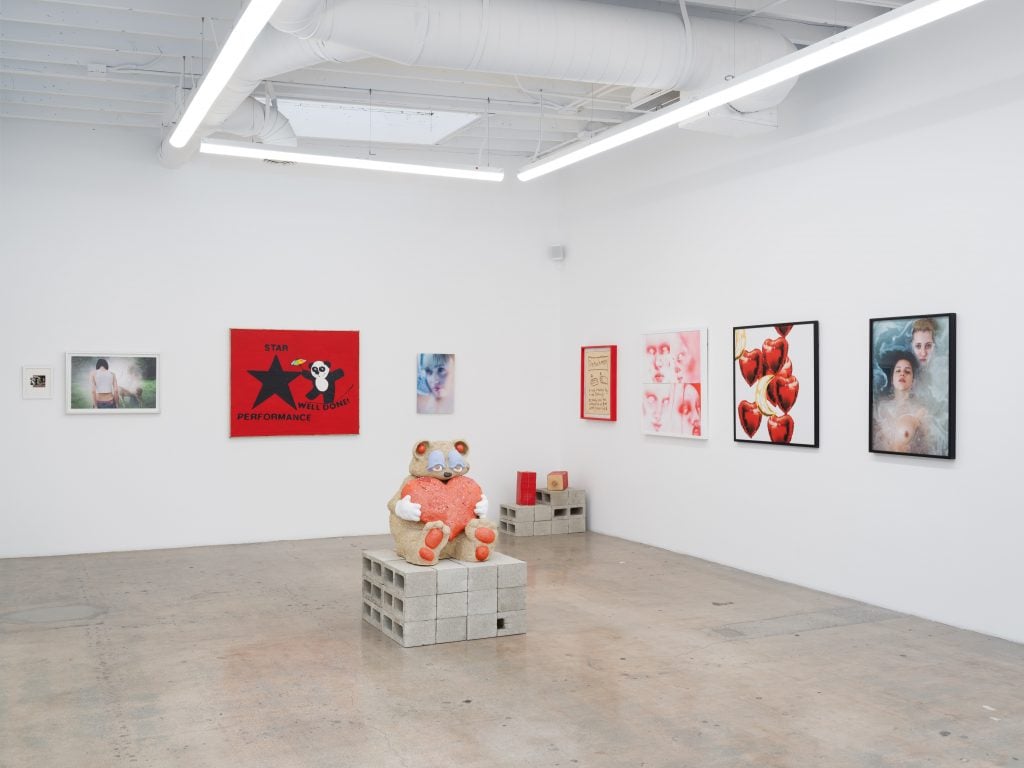 A photograph of a white walled gallery featuring punchy wall hanging artworks surrounding a sculpture of a bear holding a heart set atop a pinth of stacked cinderblocks.