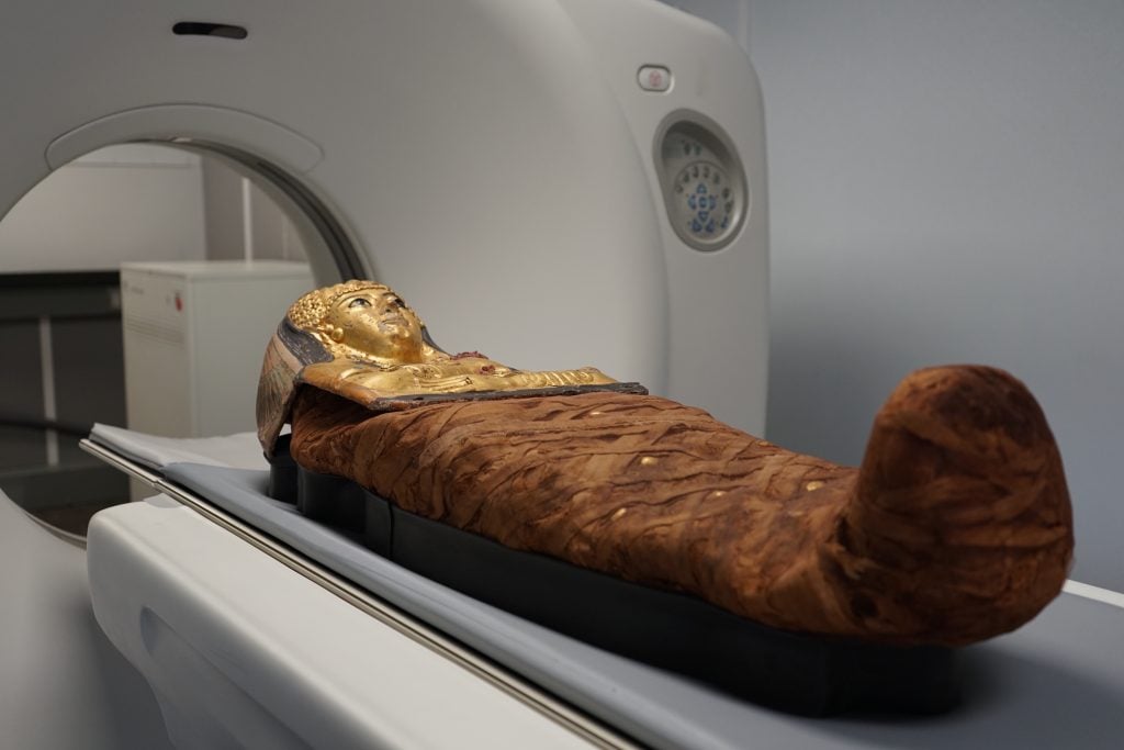 A mummy with golden deathmask and brown body wrapping lies on a CT scanner table.