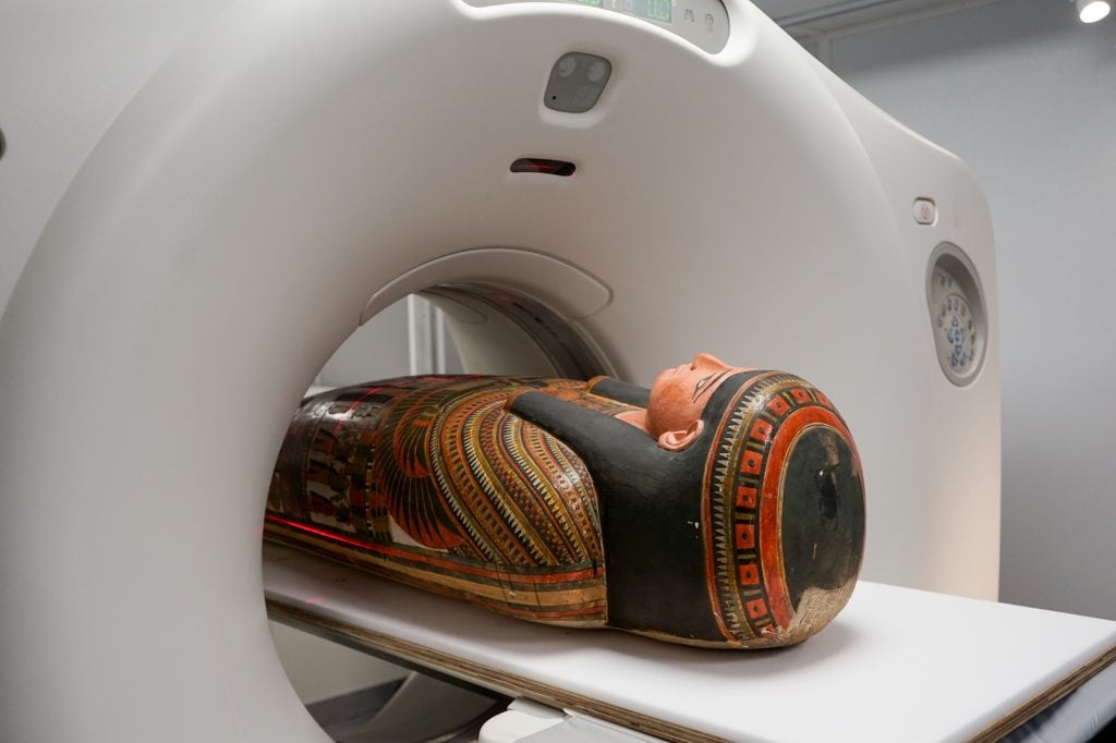 the head of an ancient mummy in a colourful casket emerges from a white CT scanner