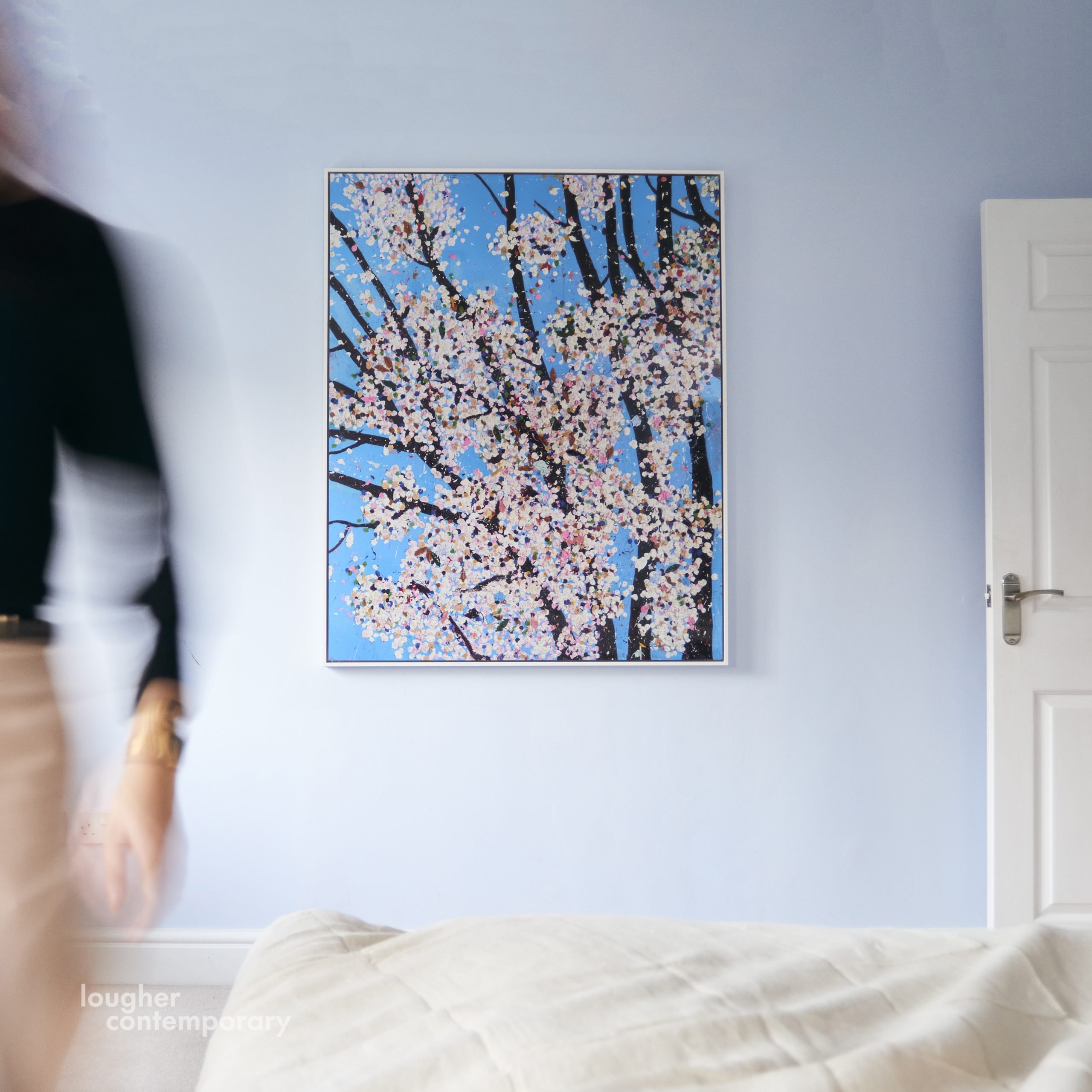 Image by Lougher Contemporary of a work by Damien Hirst of Cherry blossom tree on a pale blue wall above a bed in white linens and a blurred portion of a figure moving toward the direction of the camera.