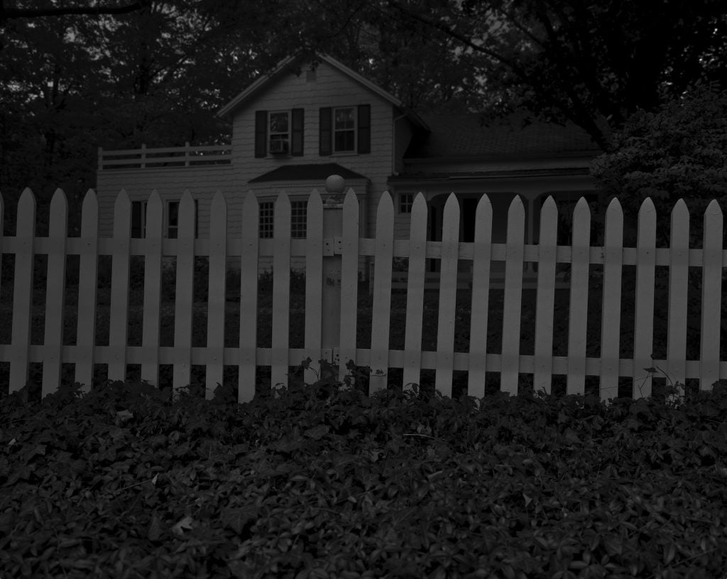  A dark, moody photograph of a house partially obscured by a white picket fence, surrounded by dense foliage and shaded trees.