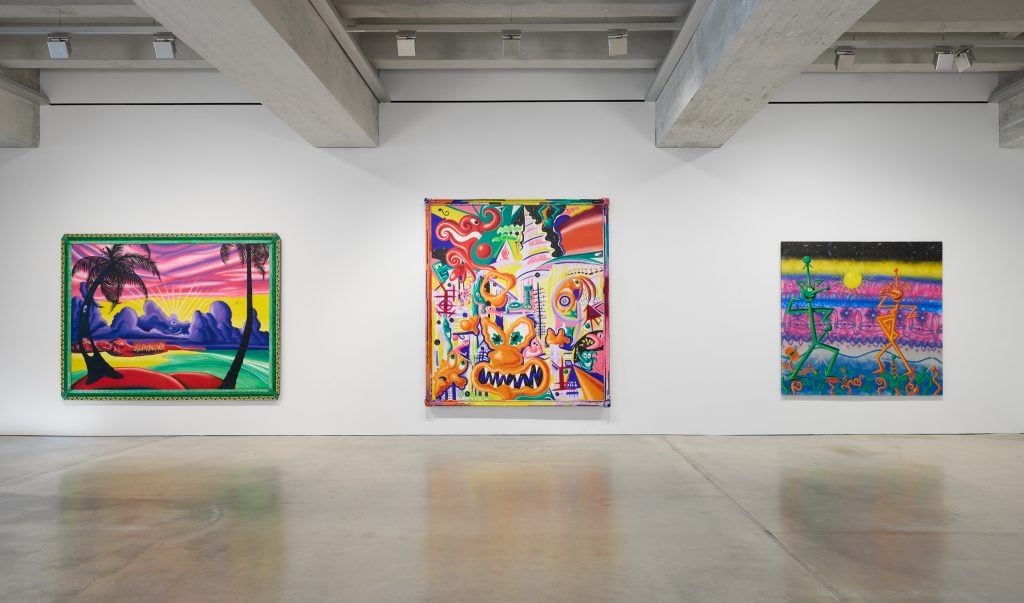 An installation photo of Kenny Scharf at the Brant Foundation, featuring three colorful paintings in a row on a white wall.