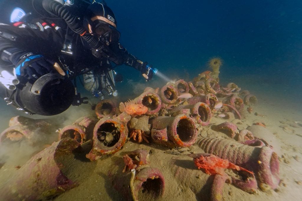 An underwater photograph looking up at a diver examinging several neat rows of Richborough Jar pottery submerged milennia ago.