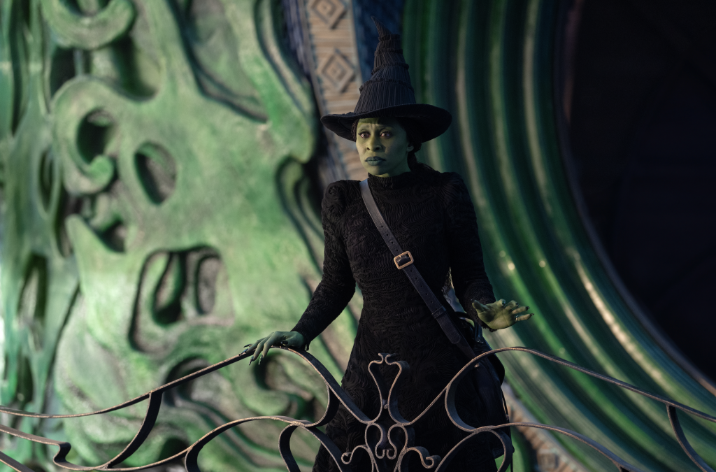 A photograph of Cynthia Erivo as Elphaba standing in a black dress and iconic black hat on the tower of the castle belonging to the Wizard.