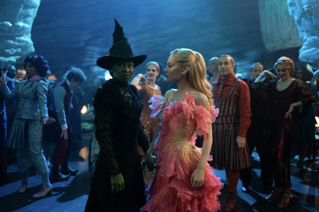 A photograph of Cynthia Erivo as Elphaba and Ariana Grande as Glinda standing in the blue-tinged Ozdust Ballroom in Wicked.